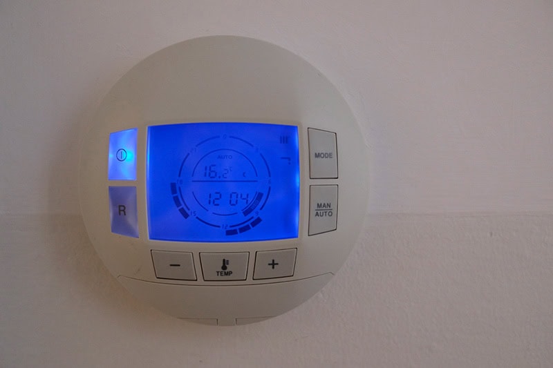 How to Set Your Programmable Thermostat. Photo of a digital thermostat mounted on a wall with a blue light.